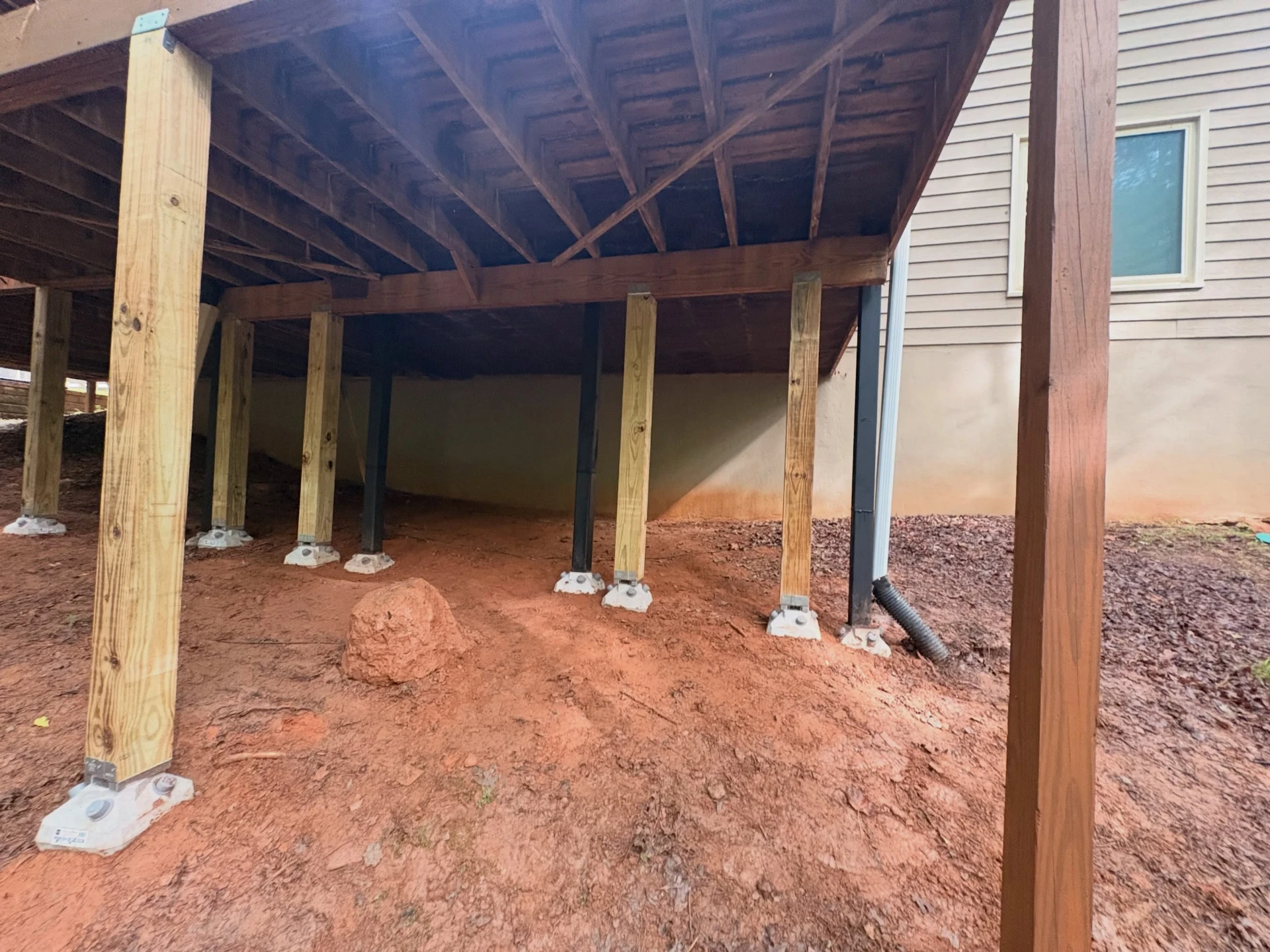 Wide view beneath a raised wooden deck showing steel support posts beside wooden posts on concrete footings