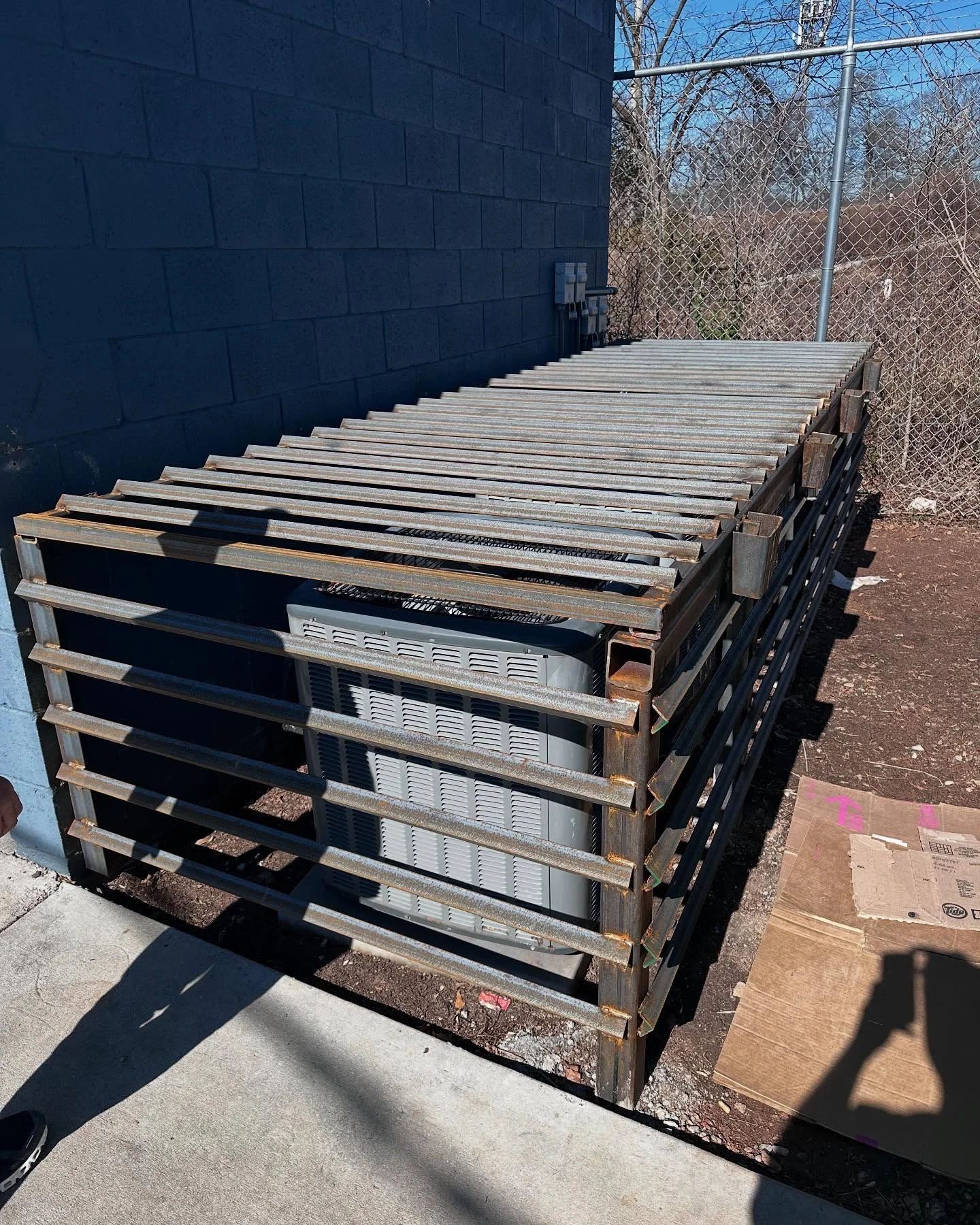 Rectangular steel security cage surrounding an outdoor equipment unit beside a block wall