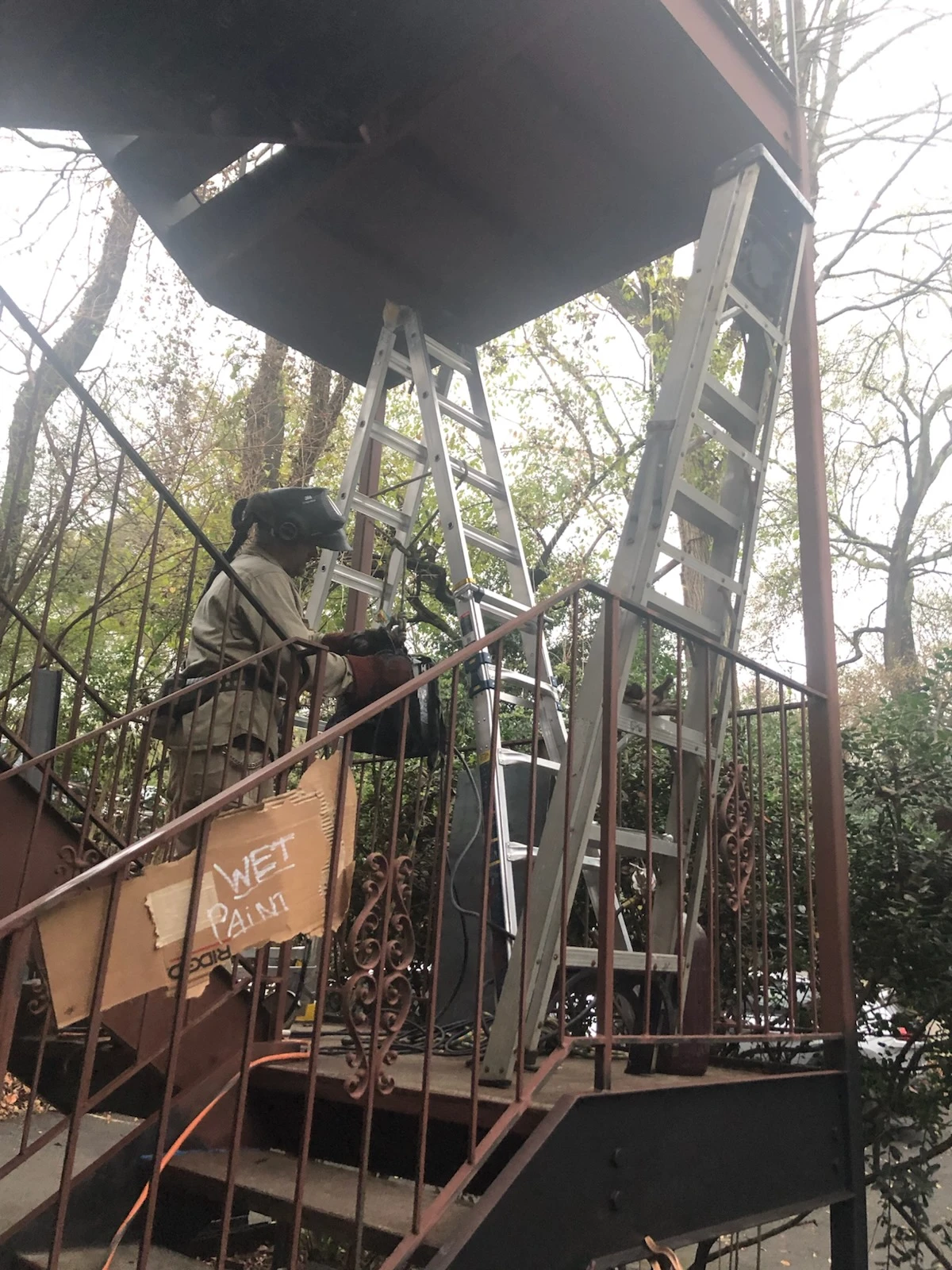 Welder working beneath an exterior steel staircase behind the stair railings