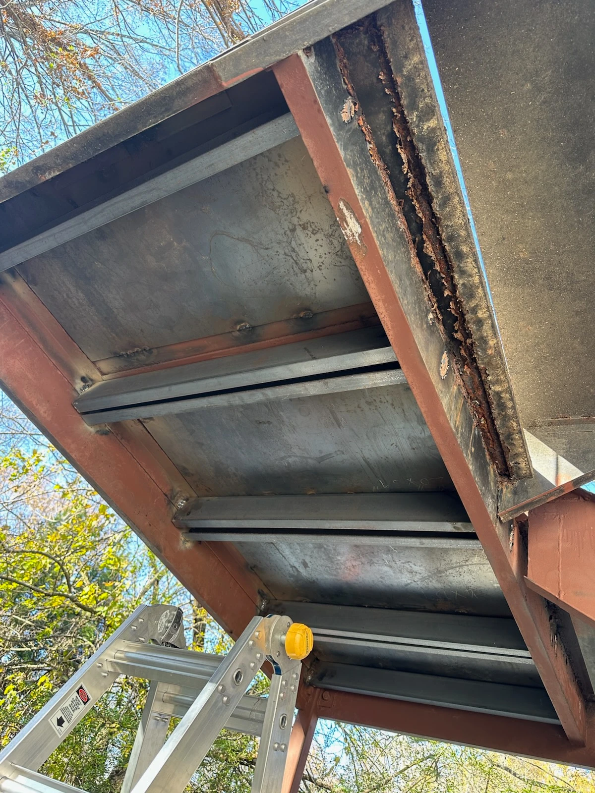Close view beneath exterior steel stairs showing rust along the tread framing and a ladder beside the staircase