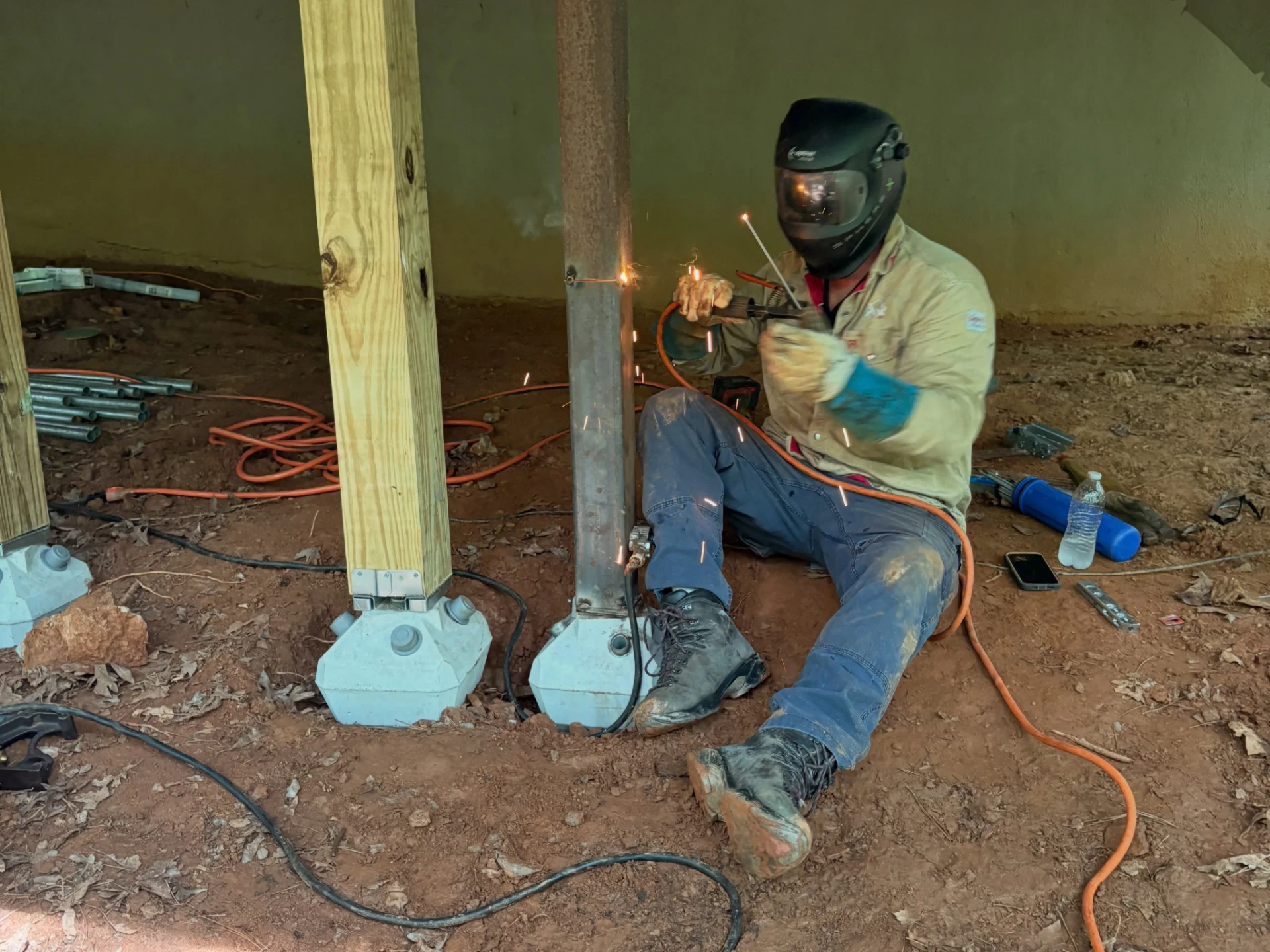 Welder kneeling beneath the deck and welding a steel support near its concrete footing